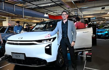 Hombre en traje claro junto a automóvil modelo S7, posando en evento de movilidad con fondo de otras personas y autos.
