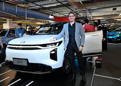 Hombre en traje claro junto a automóvil modelo S7, posando en evento de movilidad con fondo de otras personas y autos.