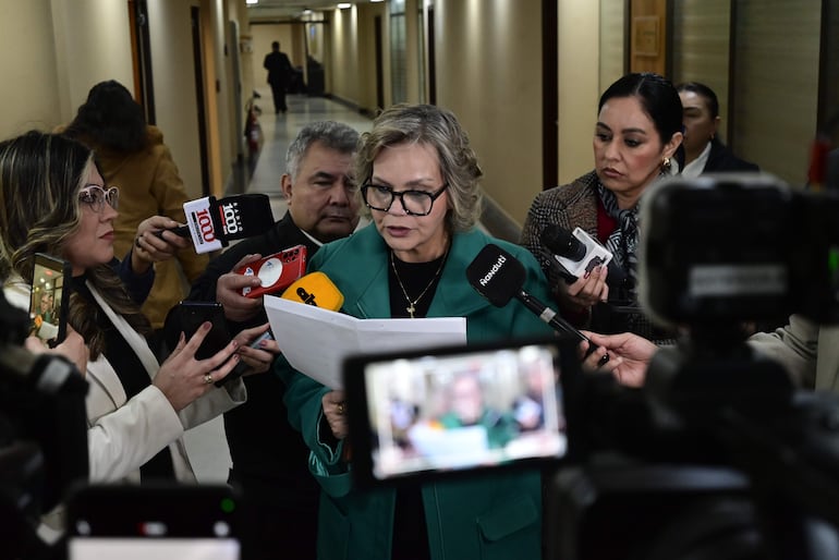 Lilian Samaniego con abrigo verde y gafas, leyendo documentos en un ambiente de oficina con reporteros y teléfonos móviles.
