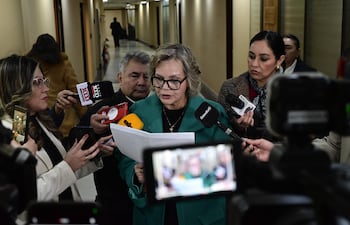 Lilian Samaniego con abrigo verde y gafas, leyendo documentos en un ambiente de oficina con reporteros y teléfonos móviles.