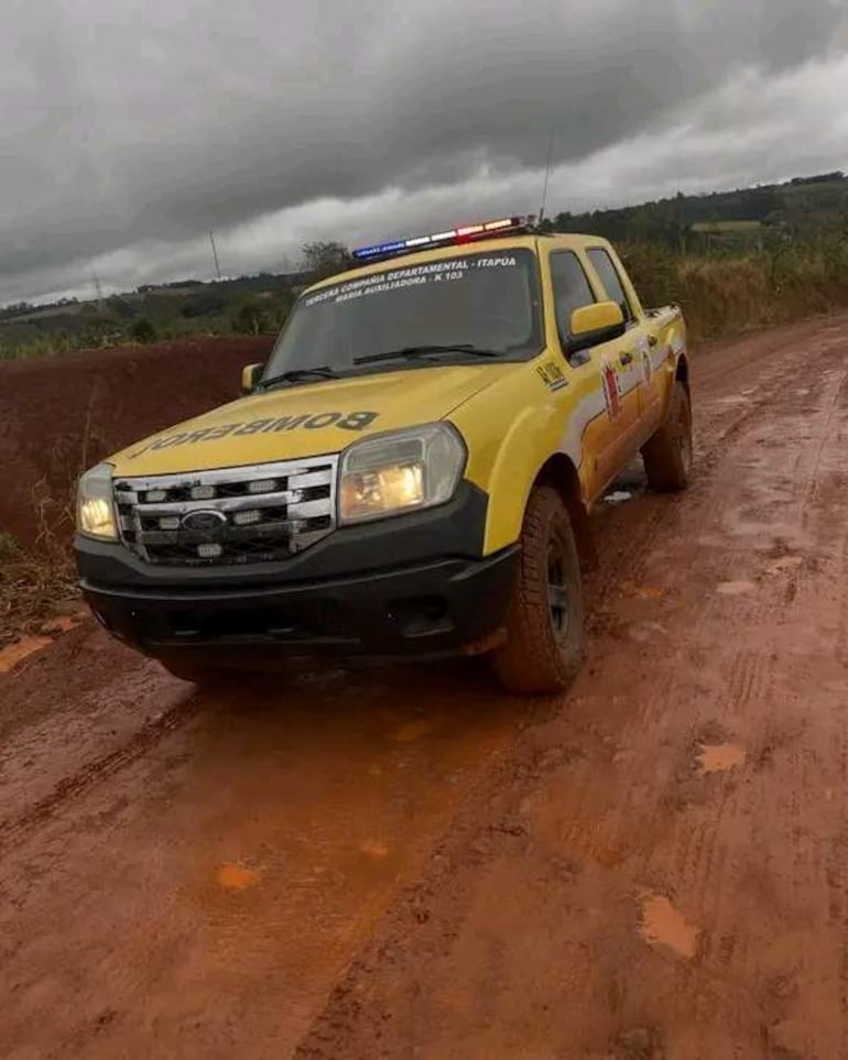 Vehículo de bomberos amarillo y negro con luces de emergencia, varado en camino fangoso rodeado de vegetación.