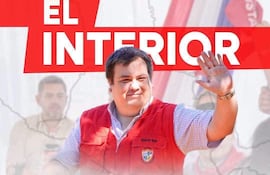 Hombre con chaleco rojo y camisa a cuadros saluda mientras otros asistentes permanecen al fondo en un evento exterior.