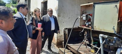 Cuatro personas conversan frente a una estructura clara; una mujer sostiene un vaso y dos hombres observan, en un ambiente de evaluación.