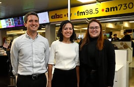 Tres personas sonrientes en una redacción, un hombre con camisa de rayas y dos mujeres, una con atuendo blanco y otra con suéter negro.