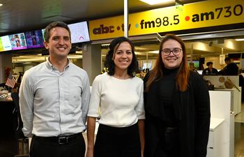 Tres personas sonrientes en una redacción, un hombre con camisa de rayas y dos mujeres, una con atuendo blanco y otra con suéter negro.
