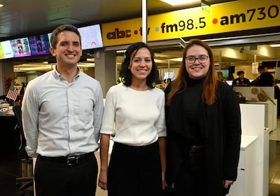 Tres personas sonrientes en una redacción, un hombre con camisa de rayas y dos mujeres, una con atuendo blanco y otra con suéter negro.