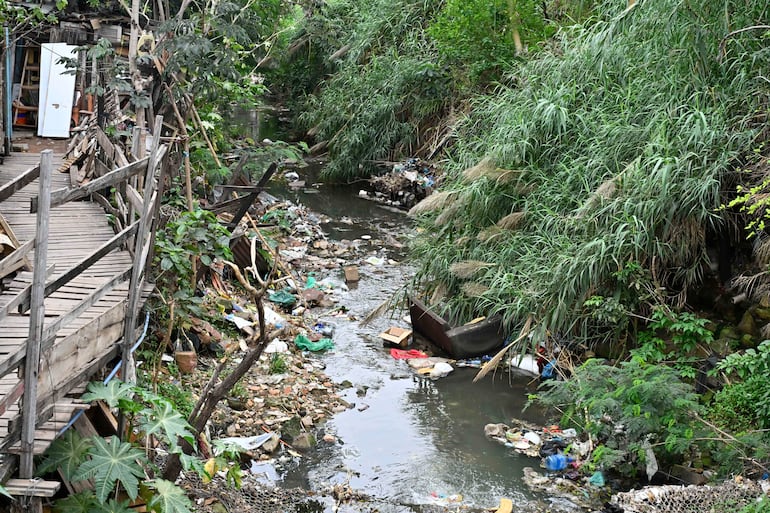 Arroyo fluido rodeado de vegetación y basura, con estructura de madera visible a la izquierda.