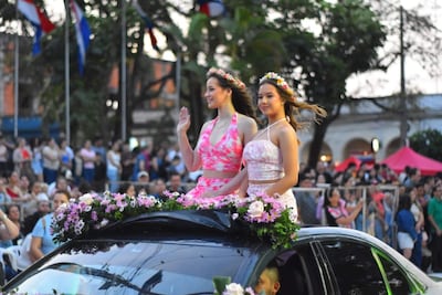 Dos mujeres en un automóvil decorado con flores, una saluda con vestido rosa y la otra observa con un vestido de tonos suaves.