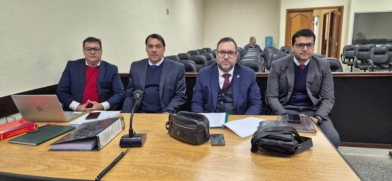 Luis Lionel Píñánez y tres hombres vestidos formalmente discuten en una sala de audiencias, con documentos y un micrófono en la mesa.
