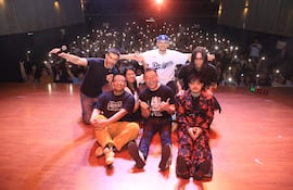 Un grupo de seis comediantes sonríe en el escenario, uno con micrófono, rodeados de luces y una audiencia entusiasta.