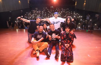 Un grupo de seis comediantes sonríe en el escenario, uno con micrófono, rodeados de luces y una audiencia entusiasta.