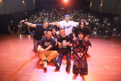 Un grupo de seis comediantes sonríe en el escenario, uno con micrófono, rodeados de luces y una audiencia entusiasta.
