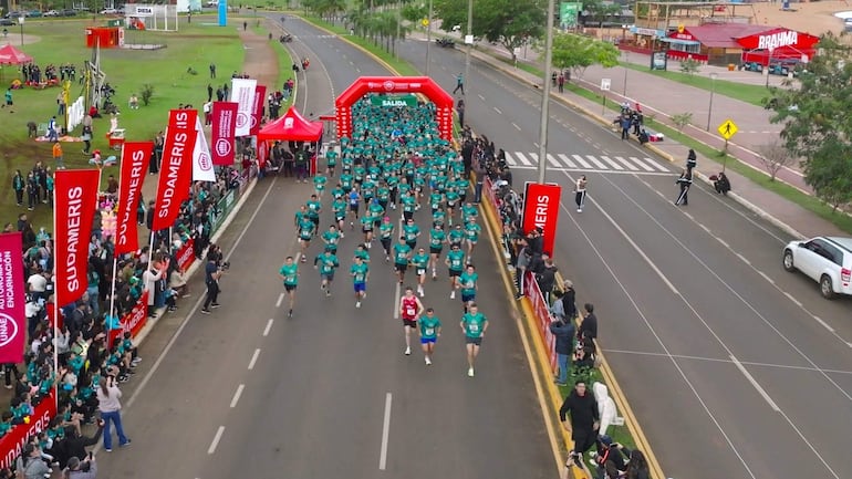 Grupo de corredores con camisetas verdes y un destacado en rojo, rodeados de espectadores animando el evento festivo.