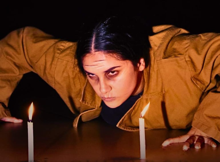 Mujer de vestido marrón arrodillada en un ambiente oscuro, concentrada en dos velas encendidas a su lado.