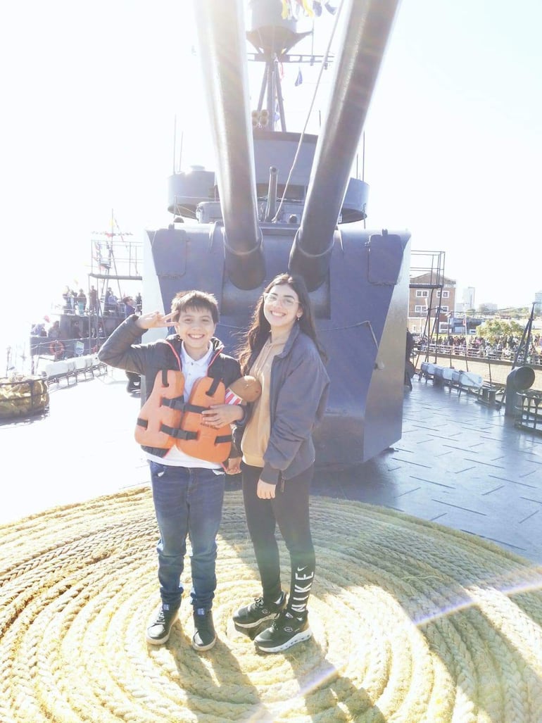 Niño con chaleco salvavidas naranja y niña en chaqueta gris sonríen en el buque, con un cañón al fondo.