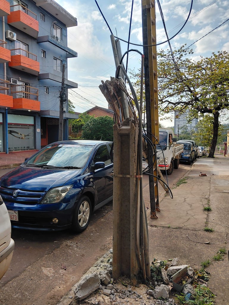 Poste de luz dañado en la calle, con cables enredados y autos estacionados en un ambiente residencial.
