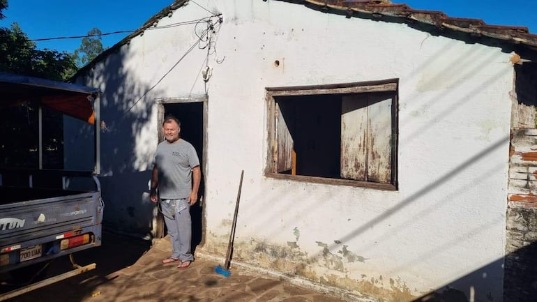 César Villalba, con camiseta gris y pantalones claros, sonríe frente a su casa de paredes blancas y techo de tejas.