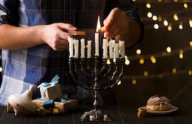 Un hombre enciende una de las velas blancas de una menorá (también conocida como januquía o candelabro de Hanukkah) en un ambiente cálido y festivo.