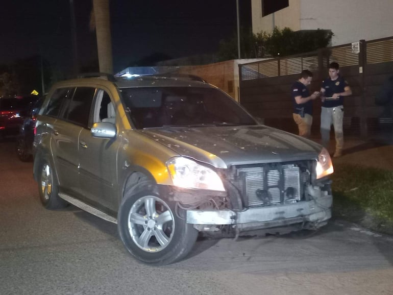 Vehículo SUV gris dañado en vía pública, dos hombres conversan con documentos en un ambiente nocturno.