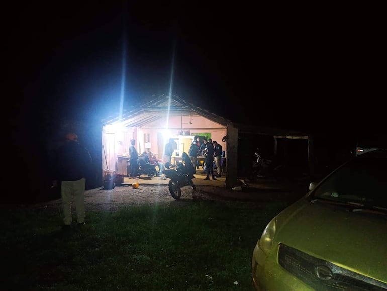 Grupo de personas observando desde fuera de una casa iluminada, con un hombre de espaldas, vestido de oscuro y con sombrero.