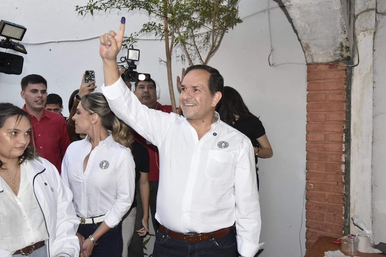 Arnaldo Samaniego sonriente con camisa blanca y cinturón marrón, rodeado de personas, mostrando su dedo con tinta azul.