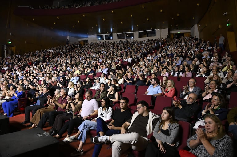 Asistentes aplaudiendo en el teatro, con vestimenta variada y gestos de emoción y celebración.