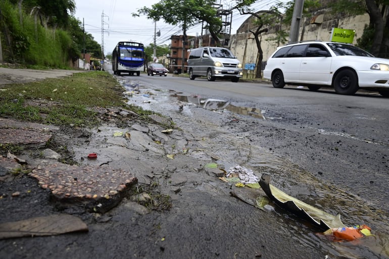 Charco de agua sucia en la acera de Avenida Artigas en Asunción, con basura y hojas esparcidas en un entorno descuidado.