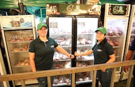 Dos mujeres en camisetas oscuras y gorras verdes sostienen puertas de refrigeradores llenos de carne en una feria.