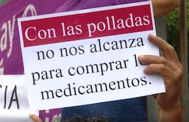 Persona sostiene un cartel que dice 'Con las polladas no nos alcanza para comprar los medicamentos', rodeada de otras personas en un ambiente urbano.