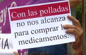 Persona sostiene un cartel que dice 'Con las polladas no nos alcanza para comprar los medicamentos', rodeada de otras personas en un ambiente urbano.