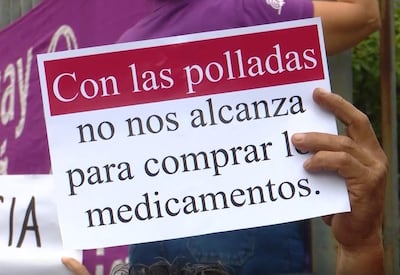 Persona sostiene un cartel que dice 'Con las polladas no nos alcanza para comprar los medicamentos', rodeada de otras personas en un ambiente urbano.