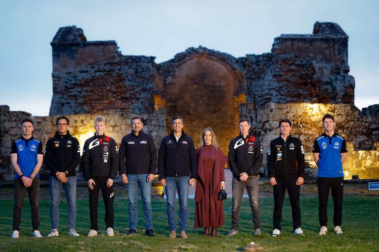 Grupo de ocho personas, algunos con chaquetas de automovilismo, posan juntos en un sitio histórico al aire libre.