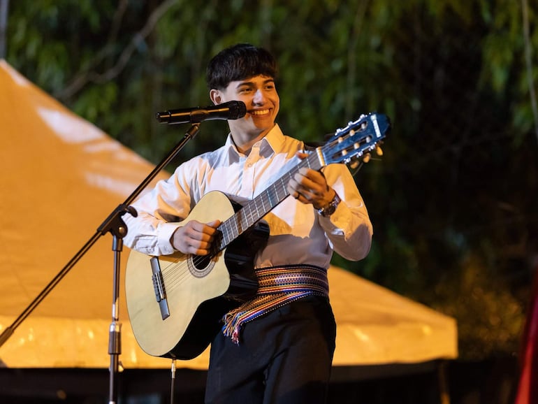 Músico sonriente con camisa blanca y cinturón tradicional, tocando guitarra al aire libre, rodeado de arbustos y carpa amarilla.