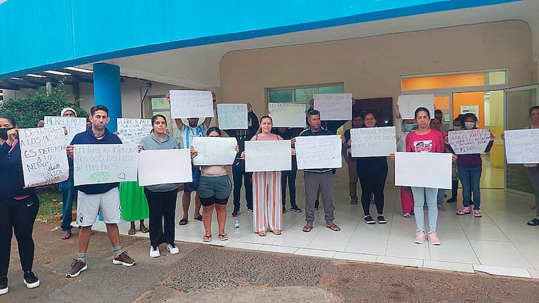 Grupo de padres con carteles en protesta, algunos visten camisetas y pantalones cortos, en ambiente exterior cerca de un edificio.