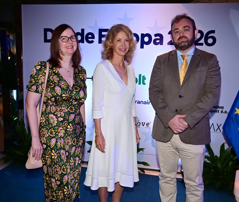 Mujer con vestido floral sonríe, otra en vestido blanco posa serena y un hombre en chaqueta gris observa con brazos cruzados.