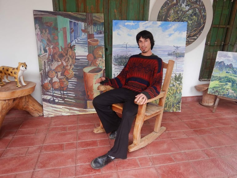 Hombre con suéter rojo y negro sentado en silla, rodeado de obras de arte en un ambiente artístico.