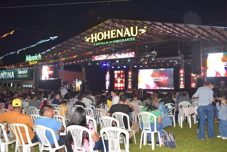 Una mujer interactúa con un niño mientras el presentador habla al público en un evento festivo con luces y decoraciones coloridas.