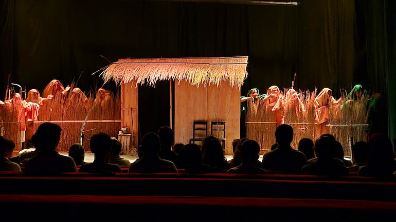 Actores en escena con vestimentas de colores terrosos, simulando una vivienda rural. Público atento en bancos rojos. Festival Internacional de Teatro en Encarnación.
