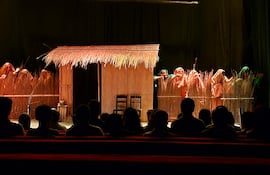 Actores en escena con vestimentas de colores terrosos, simulando una vivienda rural. Público atento en bancos rojos. Festival Internacional de Teatro en Encarnación.