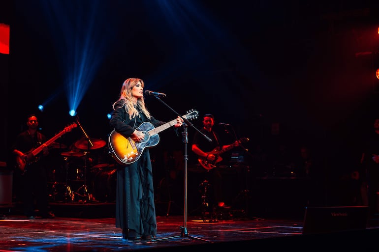 Kany García canta al micrófono con un vestido negro y guitarra acústica, acompañada de músicos en un escenario iluminado.