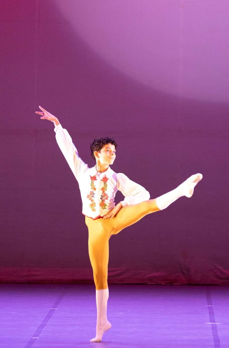 Bailarín con cabello rizado viste camisa blanca y pantalones amarillos, realizando una postura de danza en un escenario violeta.