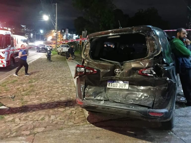 Un hombre en camiseta azul camina rápidamente mientras otro habla por teléfono, en un entorno de accidente vehicular con daños visibles.