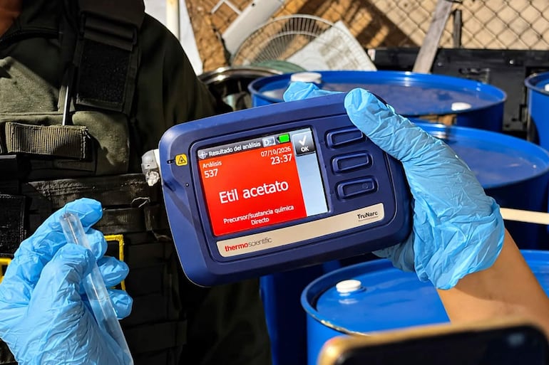 Agente con guantes azules operando dispositivo TruNarc en un entorno con barriles azules, mostrando información de análisis de acetato de etilo.