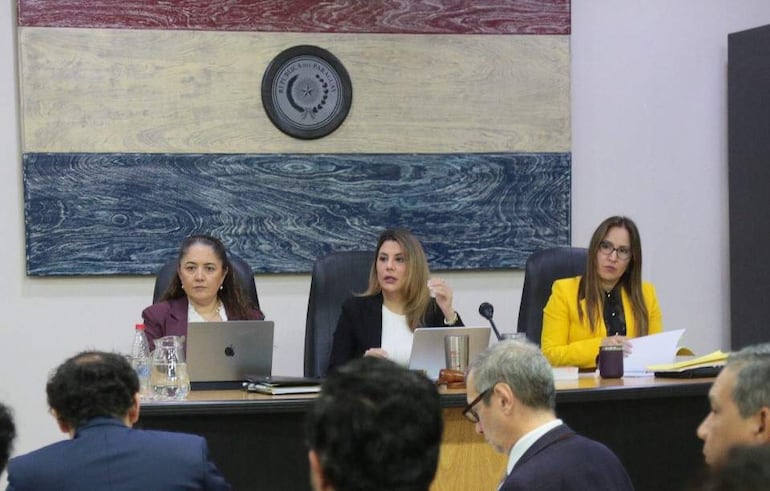 Tres mujeres en el Tribunal, una habla gesticulando, vestidas formalmente, con laptops y documentos en el escritorio. Fondo con colores de la bandera paraguaya.