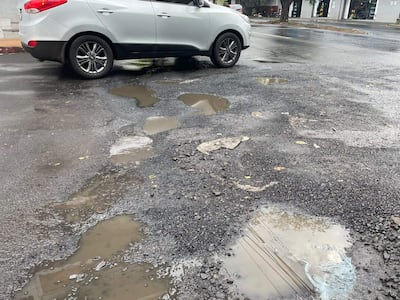 Calle asfaltada con un coche plata estacionado, baches y charcos de agua, sin personas presentes.