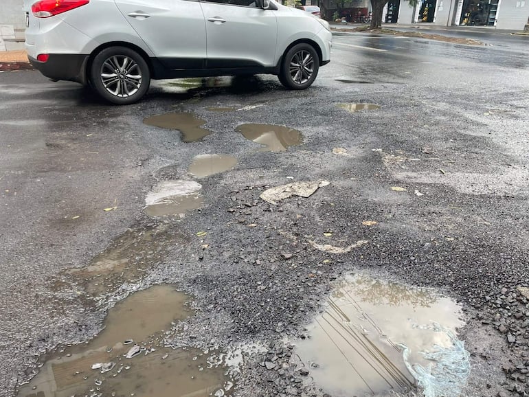 Calle asfaltada con un coche plata estacionado, baches y charcos de agua, sin personas presentes.