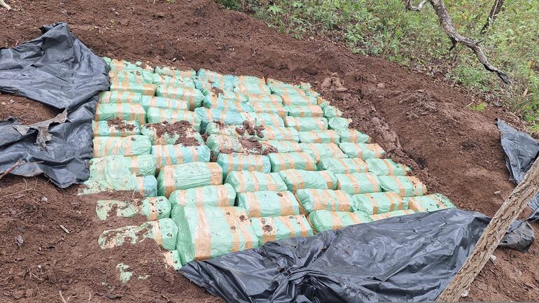 Paquetes envueltos en plástico verde y marrón apilados sobre suelo removido en un área boscosa en Paso Bravo.
