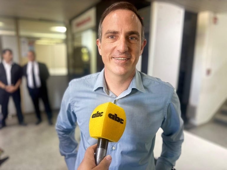 Stephan Rasmussen Gonzalez sonríe con micrófono, vestido con camisa celeste, mientras más personas observan atentamente.