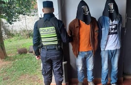Policía con uniforme observa a dos hombres encapuchados, uno con camiseta naranja y otro blanca, en un entorno verde.