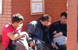 Ismael Obregón Olmedo en chaqueta oscura, sentado entre dos hombres con mascarillas, en un lugar de ladrillos al aire libre.